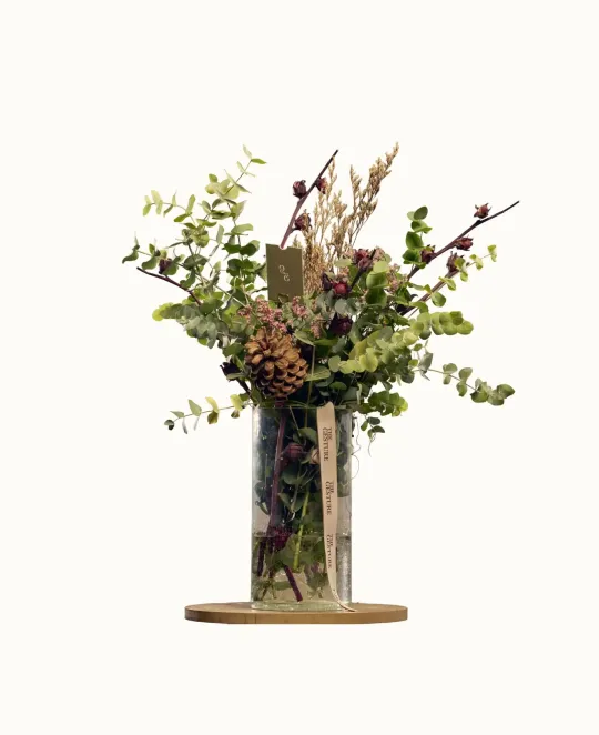 Artificial floral arrangement with greenery and branches in a clear glass vase on a wood base